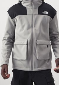 Grau Fleece-Jacke mit schwarzen Akzenten an den Schultern, zwei Fronttaschen, durchgehendem Reißverschluss und Kapuze. Der Stoff wirkt weich und strukturiert.