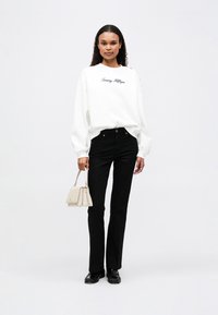 Valkoinen collegepaita, jossa on "Tommy Hilfiger" -logo, yhdistettynä mustiin räätälöityihin housuihin. Pitää kädessään pientä vaaleanbeigeä käsilaukua, jalassa mustat kengät.