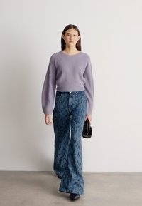 Νεαρή γυναίκα που περπατά σε εσωτερικό χώρο, φοράει λιλά πλεκτό πουλόβερ, φαρδιά μπλε τζιν με μοτίβα, μαύρα παπούτσια, κρατάει μια μικρή μαύρη τσάντα