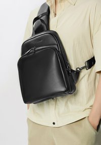Bolso de piel negro tipo bandolera con diseño cuadrado, que cuenta con un bolsillo delantero con cremallera, correa ajustable y acabado brillante.