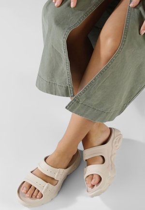 Beige sandaler med to justerbare stropper, tekstureret sål og åben tå-design bæres med en grøn, knælang beklædning med en split.
