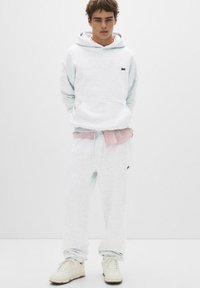 Lichtgrijze hoodie en joggingbroek van zacht materiaal, met een klein donker logo op de borst en een bijpassende grijze trekkoord in de taille.