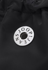 Tissu en nylon noir avec un logo circulaire argenté portant le texte "JOOP! JEANS" et une découpe centrale. Texture lisse, design minimaliste.