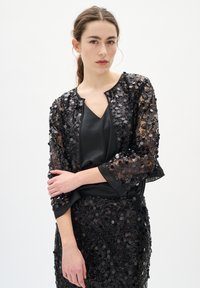 Tânără cu părul prins, purtând o jachetă și o fustă din paiete negre peste un top din satin negru, stând în fața unui fundal alb simplu.