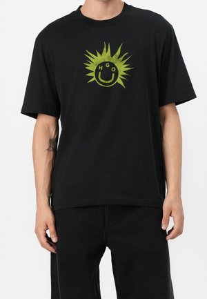 Mann, der ein schwarzes T-Shirt mit grünem Sonnenstrahl-Design und einem lächelnden Gesicht mit den Buchstaben H, G, O auf der Brust trägt, kombiniert mit schwarzen Hosen.