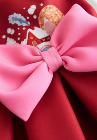 Czerwony materiał zdobi duża różowa satynowa kokarda na środku. Kokarda ma gładką fakturę, dodającą wymiaru inaczej jednolitej tkaninie.