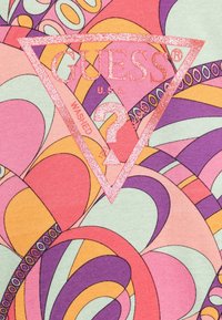 Logotipo de Guess en rosa brillante con un signo de interrogación dentro de un triángulo sobre un patrón abstracto retro colorido en tonos de rosa, púrpura, naranja y crema.