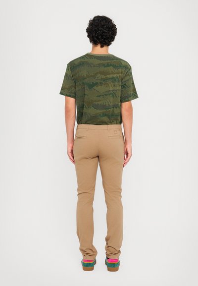 Camicia a maniche corte verde camouflage, pantaloni beige, scarpe multicolori con accenti rosa. Tessuto liscio, vestibilità rilassata, dettagli di cuciture visibili.