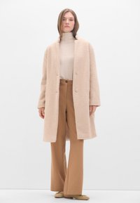 Hellbeige übergroßer Mantel aus strukturiertem, pelzähnlichem Stoff, mit großen Knöpfen, getragen über einem cremefarbenen Rollkragenpullover und braunen weit geschnittenen Hosen.