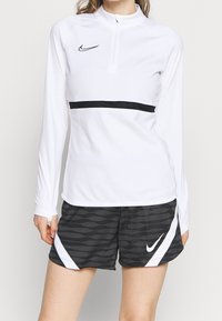 Langärmliges weißes Sportoberteil mit einem Halbzip-Kragen und einem schwarzen horizontalen Streifen, kombiniert mit schwarzen Shorts im Wellenmuster und weißen Akzenten.