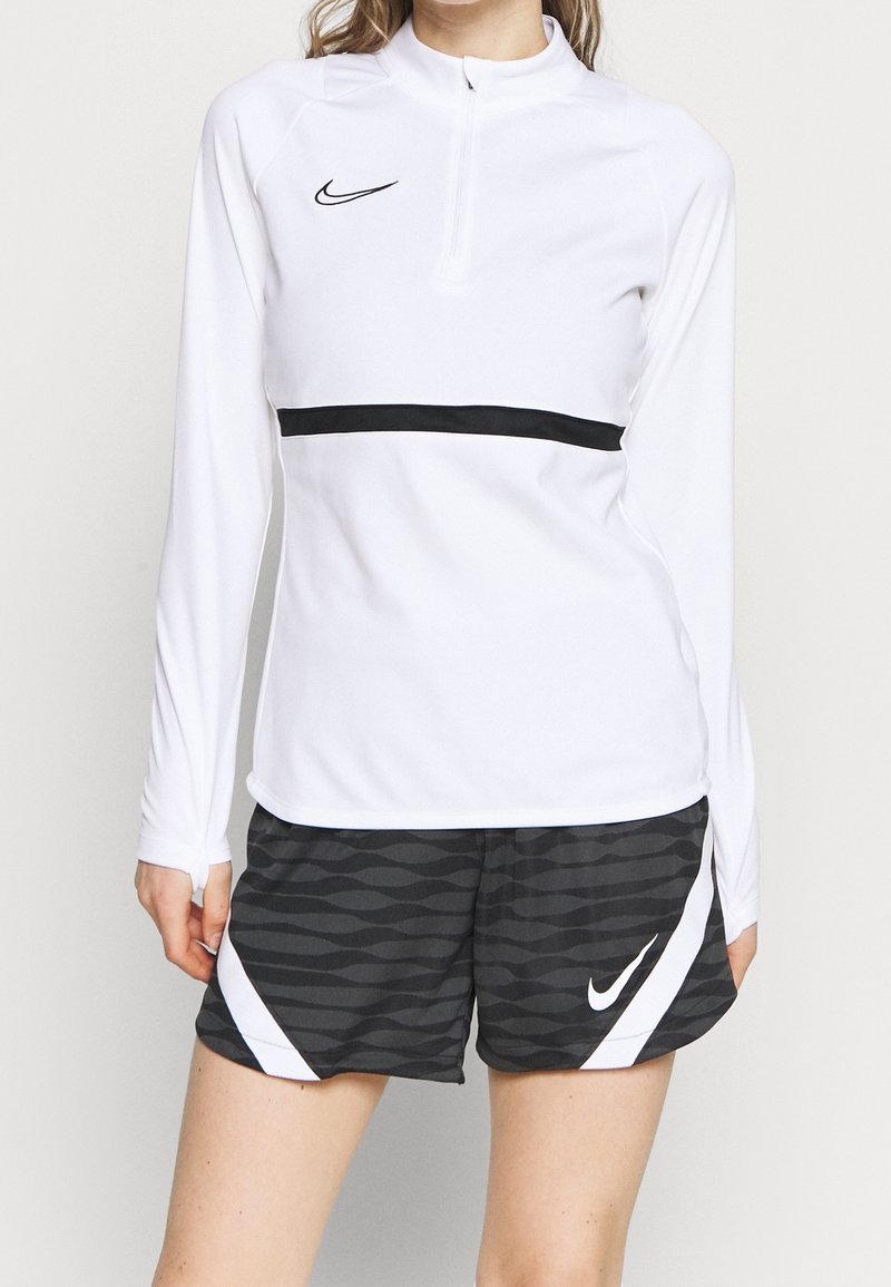 Langärmliges weißes Sportoberteil mit einem Halbzip-Kragen und einem schwarzen horizontalen Streifen, kombiniert mit schwarzen Shorts im Wellenmuster und weißen Akzenten.