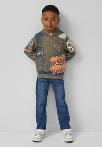 Bluza z kapturem w oliwkowym kolorze z grafikami, w tym tekstem, z przodu i na rękawach. W zestawie z niebieskimi dżinsami i białymi trampkami.