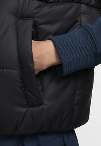 Μαύρο μπουφάν τύπου puffer με πλαϊνή τσέπη και ριμπ μανσέτα σε σκούρο μπλε χρώμα, που παρουσιάζει ομαλή υφή και ραμμένες λεπτομέρειες.