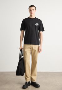 Black cotton t-shirt with graphic print, beige trousers, black tote bag, and black rubber boots, standing on a concrete floor.