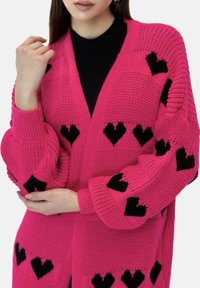Roze gebreide cardigan met zwarte hartpatronen, losse mouwen en een V-hals ontwerp. Het materiaal lijkt zacht en gestructureerd te zijn.