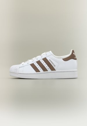 Biela teniska Adidas Superstar s hnedými pruhmi a pätkou, so škrupinovou špičkou a šnurovacím dizajnom na neutrálnom pozadí.