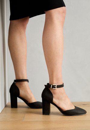 Sorte ruskind høje hæle pumps med ankelrem, lukket tå-design og en firkantet hæl. Glat tekstur, minimalistisk design og uden mønstre.