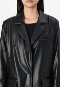 Kvinna som bär en svart fusk läderblazer över en svart topp, som är kortad vid axlarna och övre bröstet.