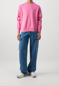 Sweatshirt rose avec col rond et poignets côtelés, associé à un pantalon cargo bleu avec des poches sur les côtés. Chaussures : baskets claires.
