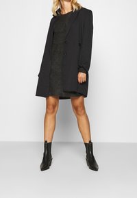 Manteau noir croisé à larges revers, associé à une robe sombre texturée. Le mannequin porte des bottines noires à bout pointu.