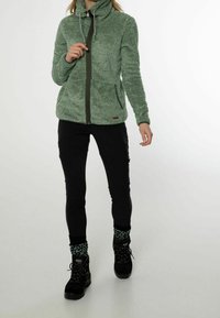 Zielona, puszysta kurtka z wysokim kołnierzem, zamkiem błyskawicznym i dwoma przednimi kieszeniami. Połączona z czarnymi spodniami i czarnymi butami. Teksturowana tkanina.