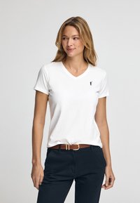Hvid bomulds t-shirt med v-hals og korte ærmer, prydet med et lille sort logo på den øverste venstre side. Bæres med mørke bukser og et brunt bælte.