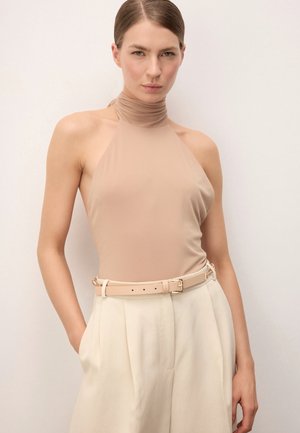 Hölgy hátul simított hajjal, ujjatlan bézs halter topban, amelyet magas derekú krémszínű nadrágba tűrt be, bézs övvel.
