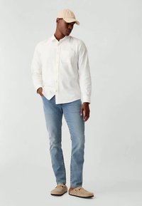 Άνδρας που φοράει μπεζ καπέλο, λευκό πουκάμισο με κουμπιά, ανοιχτό μπλε τζιν και μπεζ παπούτσια, στέκεται με το ένα χέρι στην τσέπη.