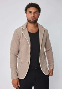 Homme aux cheveux bouclés et à la barbe, portant un blazer beige avec des bordures blanches sur une chemise noire, se tenant devant un fond gris clair.