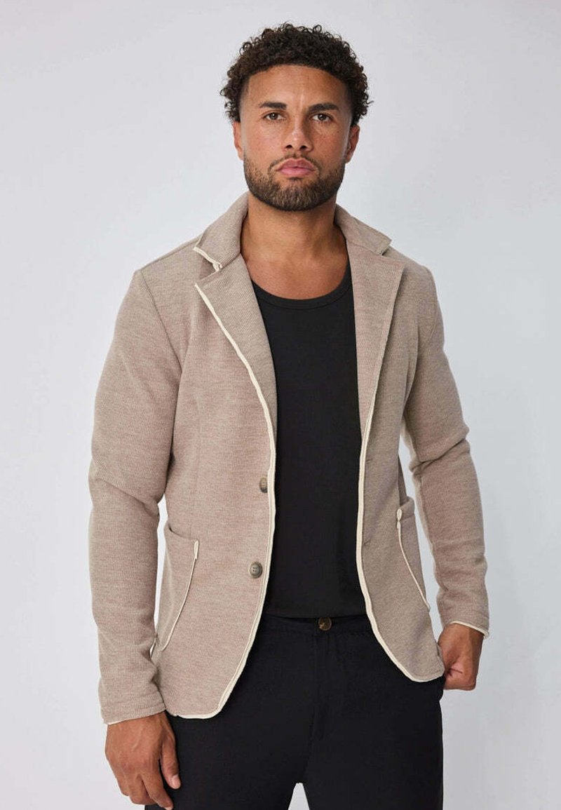 Homme aux cheveux bouclés et à la barbe, portant un blazer beige avec des bordures blanches sur une chemise noire, se tenant devant un fond gris clair.