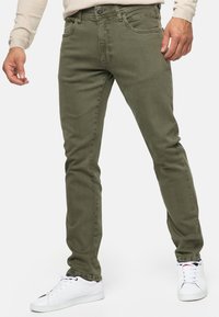 Vaqueros verde oliva de corte slim, diseño de cinco bolsillos y detalles de costura sutiles, combinados con zapatillas blancas. Textura suave y lisa.