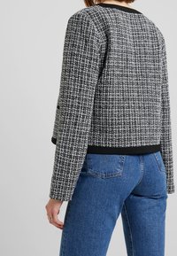 Czarna i biała kurtka z tweedu o teksturowanym, strukturalnym wzorze z czarnym, jednolitym paskiem w talii, zestawiona z niebieskimi dżinsami.