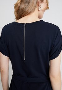 Žena v námořnickém šatu s zipem na zadní straně, krátkými rukávy a látkovým páskem, zobrazená zezadu a mírně nakloněná na stranu.