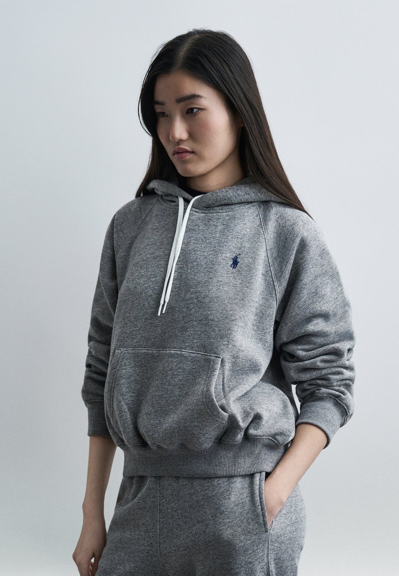 Νεαρή γυναίκα με μακριά σκουρόχρωμα μαλλιά, ντυμένη με γκρι φούτερ με κουκούλα και αντίστοιχο παντελόνι φόρμας, στέκεται με το ένα χέρι στην τσέπη μπροστά σε απλό φόντο.