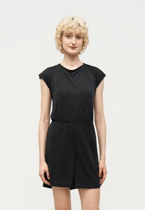 Jeune personne aux cheveux blonds bouclés et courts, portant une combinaison noire sans manches, debout devant un fond clair uni.