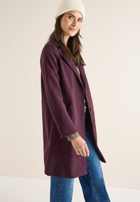 Cappotto in misto lana color bordeaux con colletto a punta, due tasche frontali e chiusura a un bottone, abbinato a un maglione grigio e jeans blu.
