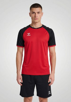 Mężczyzna stojący, ubrany w czerwoną i czarną sportową koszulkę marki Hummel oraz czarne spodenki Hummel na jednolitym jasnym tle.