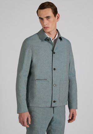 Mann trägt hellblaue, geknöpfte Jacke mit zwei Fronttaschen und passende Hose vor schlichten grauem Hintergrund.