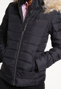 Czarna kurtka puchowa o pikowanym wzorze, z zapięciem na zamek i sztucznym futrem przy kapturze. Mały akcent logo na klatce piersiowej. Trwały materiał.