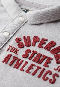 Grijze polo met een textuurstof, voorzien van een geribbelde kraag en grote rode geborduurde tekst die "SUPER TOK. STATE ATHLETICS." luidt.