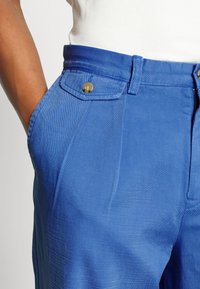 Blå bomullbyxor med en mjuk textur, med framfickor, knappstängning och subtila veck vid midjan.