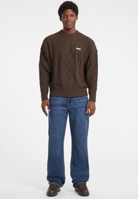 Dunkelbrauner, kabelbestrickter Pullover mit rundem Ausschnitt und gerippten Bündchen, kombiniert mit lässigen, blauem Denim-Jeans und braunen Schuhen.