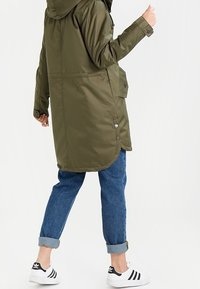 Parka imperméable vert olive avec capuche, dotée de boutons, de poches latérales et d'un dos légèrement plus long ; associée à un jean bleu avec ourlets et des baskets blanches.