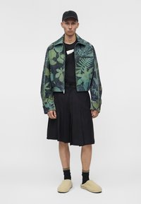 Man som bär en grön och svart mönstrad jacka, svarta plisserade shorts, svarta strumpor, beige tofflor och en svart keps, ståendes mot en enfärgad bakgrund.