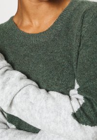 Maglione lavorato a maglia con un corpo verde, maniche grigie e colletto a coste; texture morbida e blocchi di colore a contrasto riconosciuti.