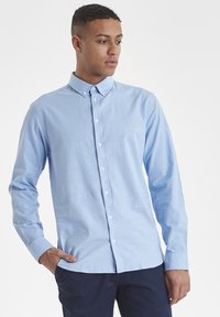 Camisa azul claro de manga larga con botones, confeccionada en algodón. Presenta un cuello clásico, botones en el frente y un logo bordado sutil en el pecho.