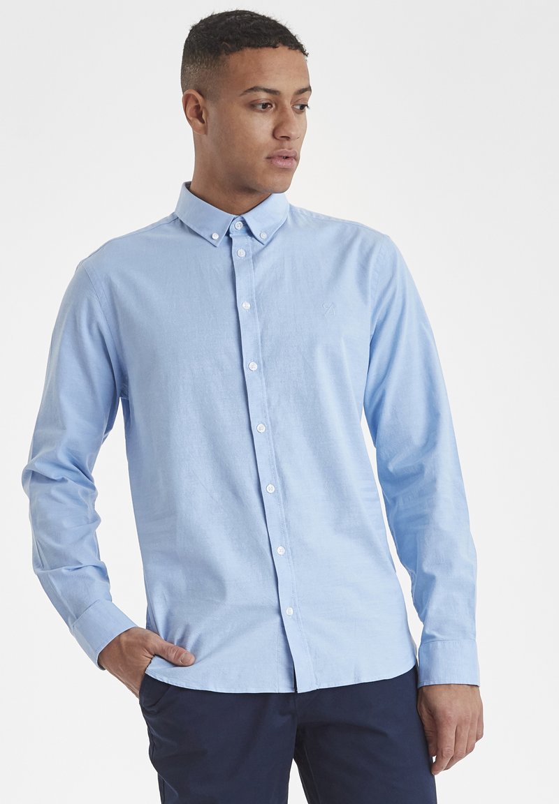 Camisa azul claro de manga larga con botones, confeccionada en algodón. Presenta un cuello clásico, botones en el frente y un logo bordado sutil en el pecho.