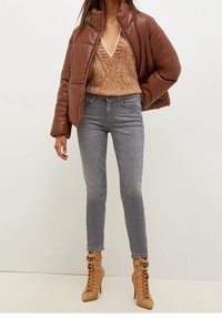 Brun polstret jakke, strikket beige top, grå slim-fit jeans og beige støvler med høje hæle, snøredetalje og gulddetaljer.