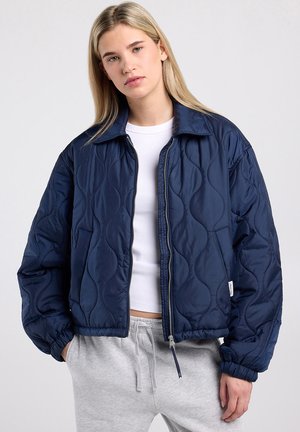 Jonge vrouw met steil blond haar, gekleed in een marineblauwe doorgestikte jas, een wit cropped shirt en grijze joggingbroek met handen in de zakken.