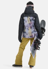 Snowboarder in a black and pastel mountain-patterned jacket, yellow pants, and black boots, holding a snowboard with bindings.